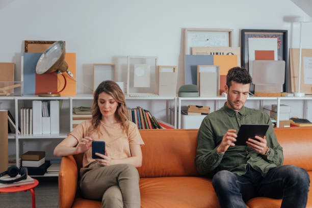 Image de deux personnes assises chacune d'un bord du sofa ayant en main et consultant un cellulaire et une tablette